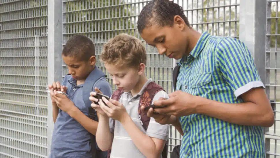 Three boys are standing along a fence looking at their phones.