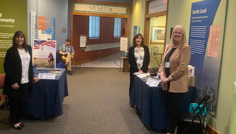 Three women stand behind informational tables inside a museum exhibit hallway, smiling toward the camera. Display boards with text and images are set up around them, including one titled ‘Fertile Land.’ A man sits in the background near the entrance labeled ‘Museum'