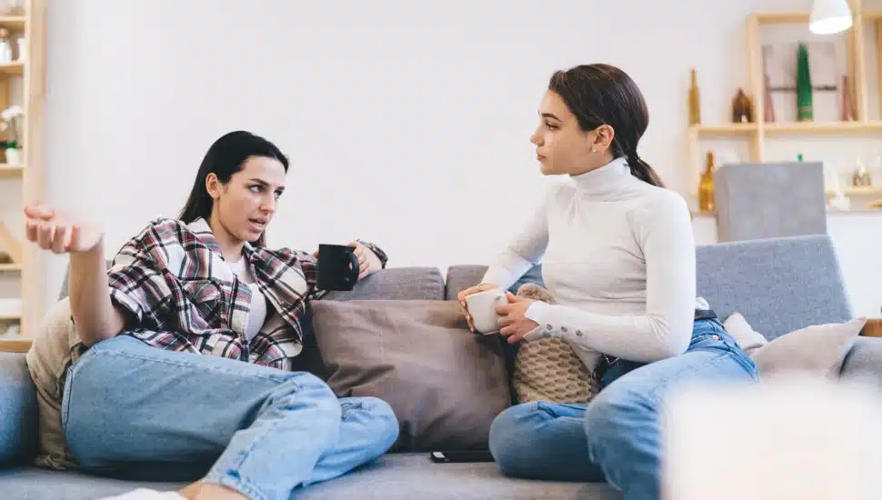 Two women sit on a couch. One speaks as the other listens.