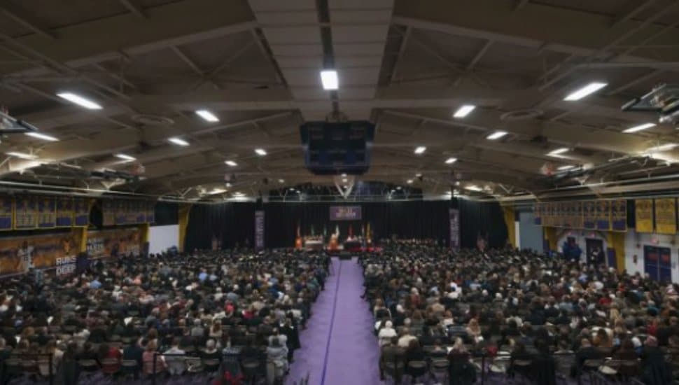 Hollinger Field House filled with hundreds of guests and graduates during a West Chester University commencement ceremony, with a stage and banners visible at the front of the arena.