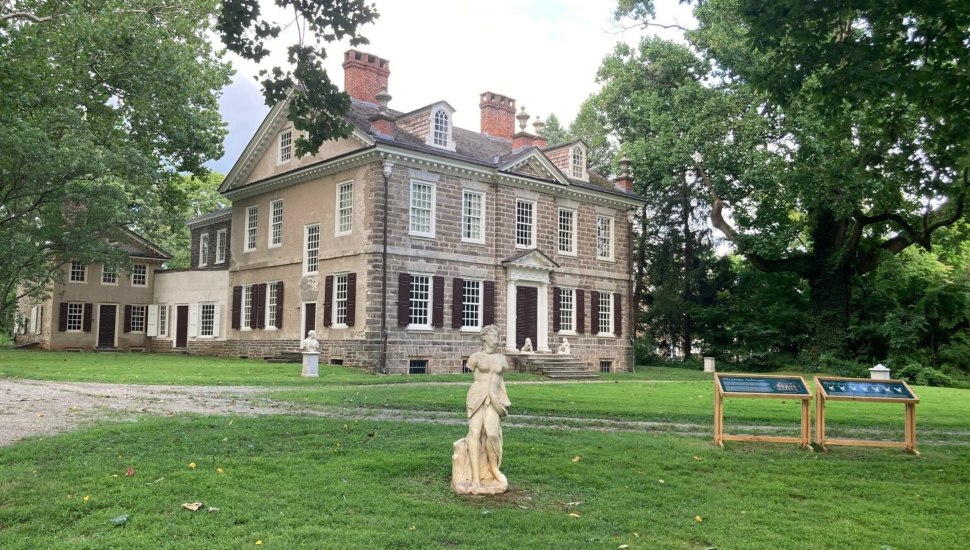 Exterior view of Cliveden, a two-story Georgian stone mansion in Germantown, Philadelphia, with classical statues on the lawn and mature trees surrounding the property.