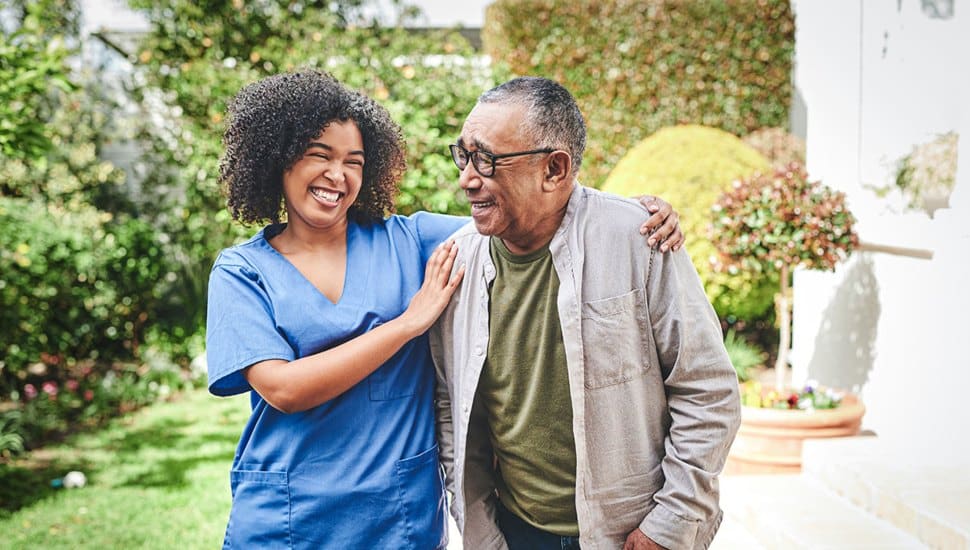 A health care worker walks side by side with an older man who is using a cane to walk.