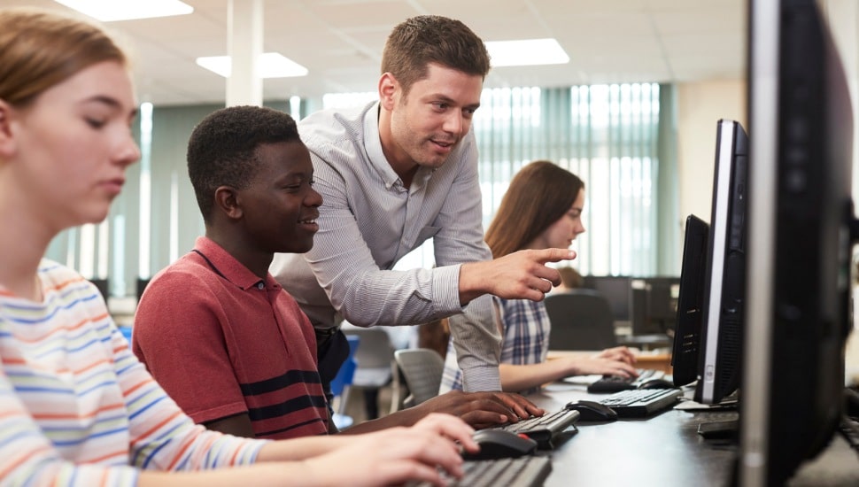 High school students work at computers, assisted by a teacher.