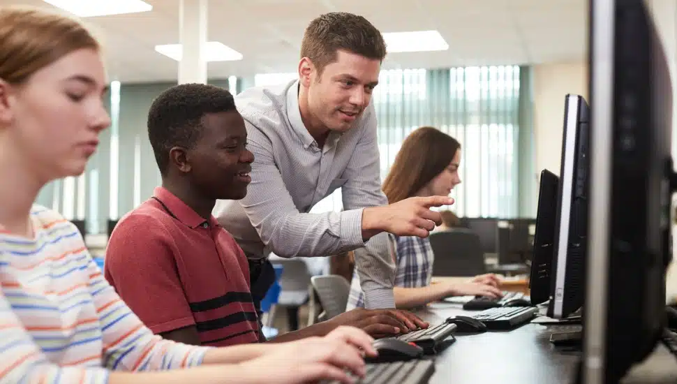 High school students work at computers, assisted by a teacher.